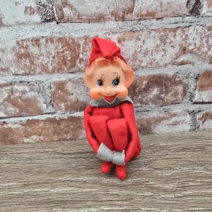 May include: A vintage Christmas elf doll with a red felt outfit, hat, and shoes. The elf has a plastic face with rosy cheeks, blue eyes, and a gray collar. The doll is sitting on a wooden surface in front of a brick wall.