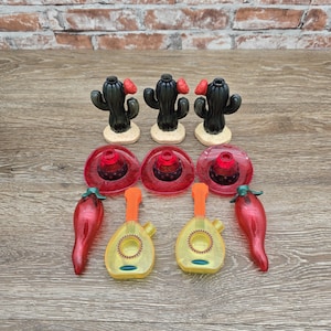 May include: A collection of decorative items with a Mexican theme. The set includes three black cactus figurines with red flowers, three red sombrero-shaped pieces, two yellow mandolins, and two red chili pepper figurines. The items are arranged on a wooden surface.