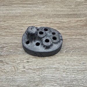 May include: A dark gray, circular object with multiple holes and a textured surface. The object has a central, raised element and appears to be made of a solid material. The item is likely a decorative or functional piece.