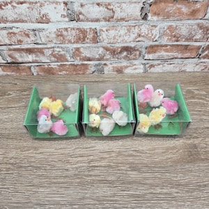 May include: Three clear plastic containers, each holding several small, fluffy chicks in shades of pink, yellow, and white. The chicks are arranged on a green base, with a brick wall in the background. The overall theme is Easter or spring.