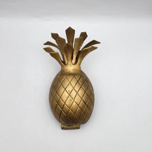 May include: A gold-colored pineapple door knocker with a detailed design. The knocker is made of metal and has a realistic pineapple shape.