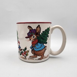 May include: A white ceramic mug with a red rim features a Christmas-themed illustration of a brown rabbit wearing a blue jacket and a striped hat, carrying a small green Christmas tree. The rabbit is surrounded by holly, berries, and a small bird.