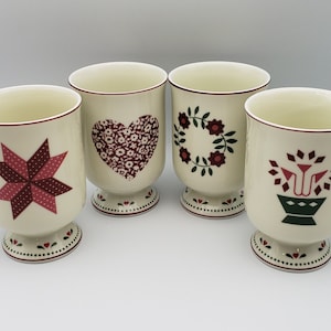 May include: Four cream-colored ceramic mugs with decorative designs. Each mug features a different design: a star, a heart, a floral wreath, and a floral basket. The mugs have a footed base and a handle, with a red trim.