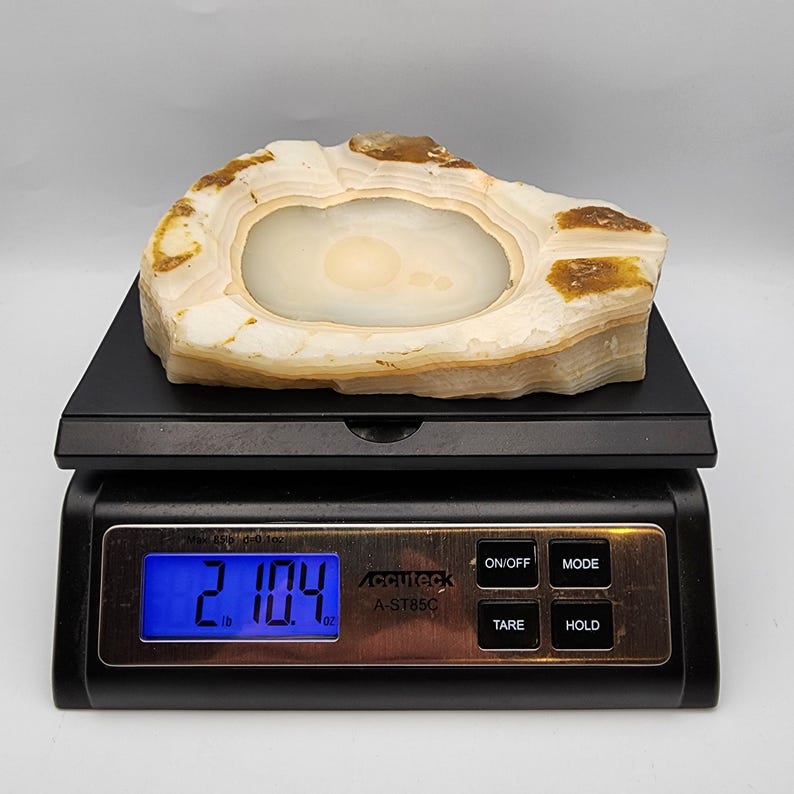 May include: A natural stone bowl with a central depression, resting on a black digital scale. The bowl is off-white with brown and tan banding. The scale displays 2.104 lb.