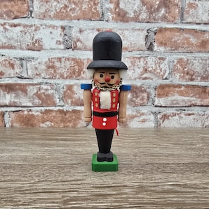 May include: A wooden nutcracker figurine with a black hat, red jacket, and black pants. The figurine has a gold chain around its neck and is standing on a green base. The background is a brick wall.