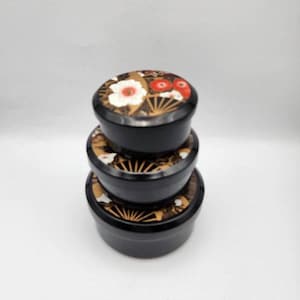 May include: A stack of three round black containers with lids. Each lid features a decorative pattern of gold fan shapes, white flowers, and small red circular accents. The containers are arranged in descending order of size from bottom to top.