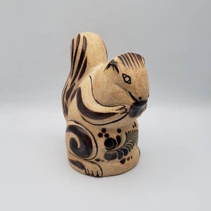 May include: A ceramic squirrel figurine in a seated pose, holding a dark object. The squirrel has a tan body with dark brown stripes and decorative floral patterns. The figurine has a glossy finish and is set against a plain background.