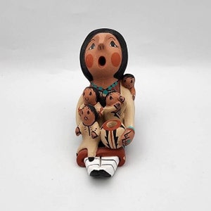 May include: A handcrafted ceramic storyteller figurine. The figure is tan with black hair and a wide open mouth. It is surrounded by four smaller figures and holds a patterned pot. The base is red and white.
