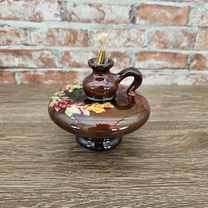 May include: A brown ceramic oil lamp with a glossy finish, featuring a small handle and a central wick holder. The base is decorated with a raised, colorful floral pattern in shades of green, yellow, and red. The lamp has a rounded, disc-shaped body.