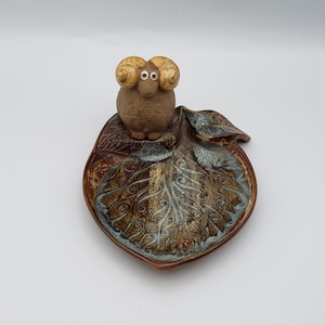 May include: A ceramic ram figurine with curled horns sits atop a leaf-shaped dish. The dish features a detailed leaf vein design with brown, gold, and blue-gray hues. The ram is brown with tan horns and white eyes.