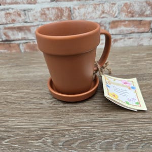 May include: A terracotta-colored mug shaped like a flower pot, with a matching saucer. The mug has a handle and a tag tied to it with twine. The tag has text and floral illustrations.