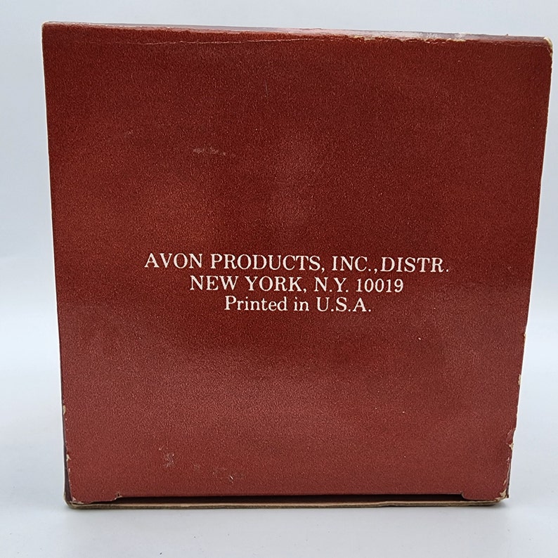 May include: A rectangular, reddish-brown box with white text that reads "AVON PRODUCTS, INC., DISTR. NEW YORK, N.Y. 10019 Printed in U.S.A." The box has a slightly worn appearance.