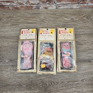 May include: Three vintage Christmas ornaments in their original packaging. Each ornament is a whimsical figure with a unique design, including a pink-faced character with a long white beard, a character with a yellow and black outfit, and a character with a red and blue outfit.