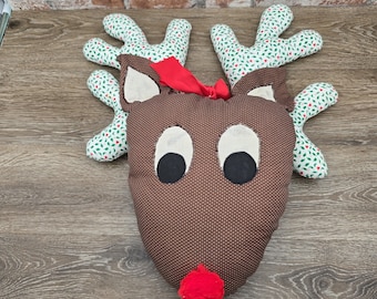 May include: A handmade reindeer head-shaped pillow with a brown polka-dot face, large black eyes, and a red pom-pom nose. The antlers are green with a white and red floral pattern, and a red bow is tied between the antlers. A festive holiday decoration.