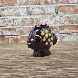 May include: A ceramic fish figurine with a dark brown glaze and yellow polka dots. The fish has a detailed fin and tail, and a small, round eye. The figurine is displayed on a wooden surface against a brick wall.