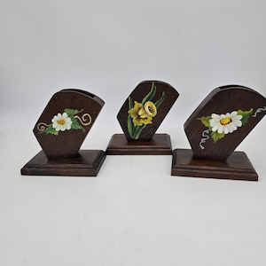 May include: Three dark brown wooden napkin holders with rectangular bases. Each holder features a hand-painted floral design: two display white daisies with green leaves, and the center holder features a yellow daffodil with green stems.