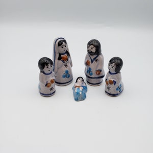 May include: A set of five hand-painted ceramic figurines featuring a central infant, two taller adult figures, and two smaller figures. Each piece is light gray with black hair, blue floral accents, and brown circular details on the clothing.