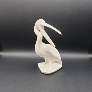 May include: White porcelain figurine of a pelican with its beak curved over its head. The pelican is standing on a base with a textured design.