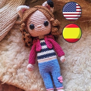 Op de afbeelding: Een gehaakte pop met bruin haar, die een roze jasje, een gestreept shirt, een blauwe spijkerbroek en roze schoenen draagt. De pop heeft grote zwarte ogen en een roze neus. De pop zit op een pluche wit oppervlak met een Amerikaanse vlag en een Spaanse vlag op de achtergrond.