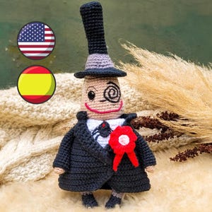 Może przedstawiać: Pluszowa zabawka wykonana na szydełku przedstawiająca postać w czarnym garniturze, cylindrze i czerwoną rozetą. Postać ma spiralne oko i przyjazny uśmiech.