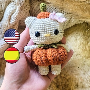 Puede incluir: Una muñeca Hello Kitty de crochet vestida de calabaza. La muñeca es blanca con ojos negros y un lazo rosa. La calabaza es naranja y tiene un tallo verde.