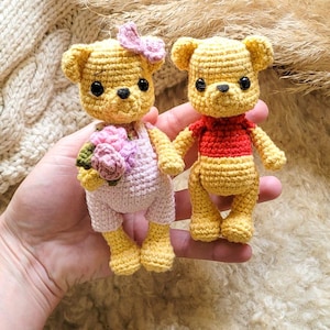 Puede incluir: Dos osos de peluche de ganchillo hechos a mano. Uno lleva un lazo rosa y un peto, sosteniendo un ramo de flores rosas. El otro oso lleva una camisa roja. Ambos son amarillos con ojos y nariz negros.