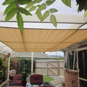 May include: A beige shade sail is stretched over a white pergola, providing shade for a patio with two red chairs and a glass table. The patio is surrounded by greenery and a wooden fence.