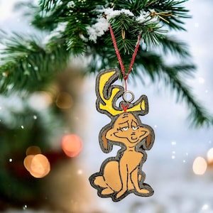 May include: A Christmas ornament featuring a cartoon dog with yellow antlers. The ornament is brown and yellow with a red string for hanging. The ornament is hanging from a snow-covered evergreen branch.