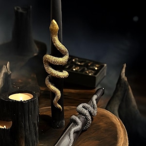 May include: Black candles adorned with snake embellishments. One candle has a gold snake, the other a silver snake. The candles are presented on a wooden surface, creating a gothic or mystical aesthetic. The candles are approximately 20 cm tall.
