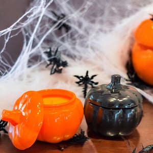 Puede incluir: Dos recipientes con forma de calabaza, uno naranja y otro negro, están sobre una superficie de madera. El recipiente naranja está abierto, revelando una vela amarilla en su interior. Arañas negras y telarañas blancas decoran el fondo.