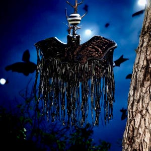 May include: A black metal bat decoration with fringe, suspended from a branch. The bat features detailed wings and a textured body. The background is a dark blue night sky with bat silhouettes.