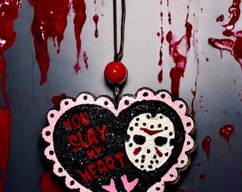 May include: A heart-shaped pendant with the text "YOU SLAY MY HEART" and a hockey mask design. The pendant is black with pink trim and a red bead at the top. Red splatters are in the background.