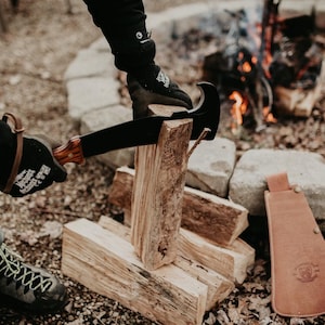 May include: A person wearing black gloves is using a black and brown machete to chop wood. The wood is stacked in a pile next to a fire pit. A brown leather sheath is on the ground next to the wood.