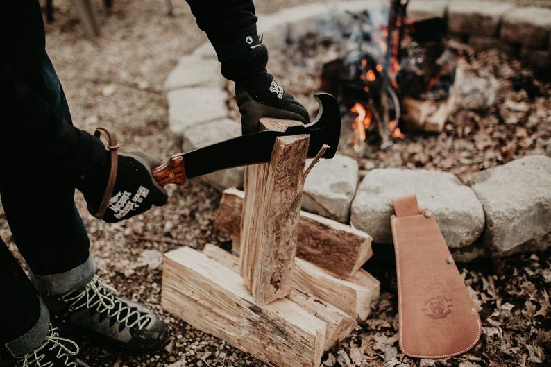 May include: A person wearing black gloves is using a black and brown machete to chop wood. The wood is stacked in a pile next to a fire pit. A brown leather sheath is on the ground next to the wood.