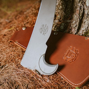 May include: A silver-colored tool with a curved blade and a leather sheath. The sheath is brown and has the text "Western Trail" embossed on it.