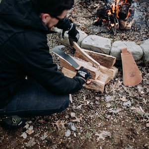 May include: A person wearing black gloves uses a large black knife to split a piece of firewood near a campfire. The knife has a curved blade and a leather sheath.