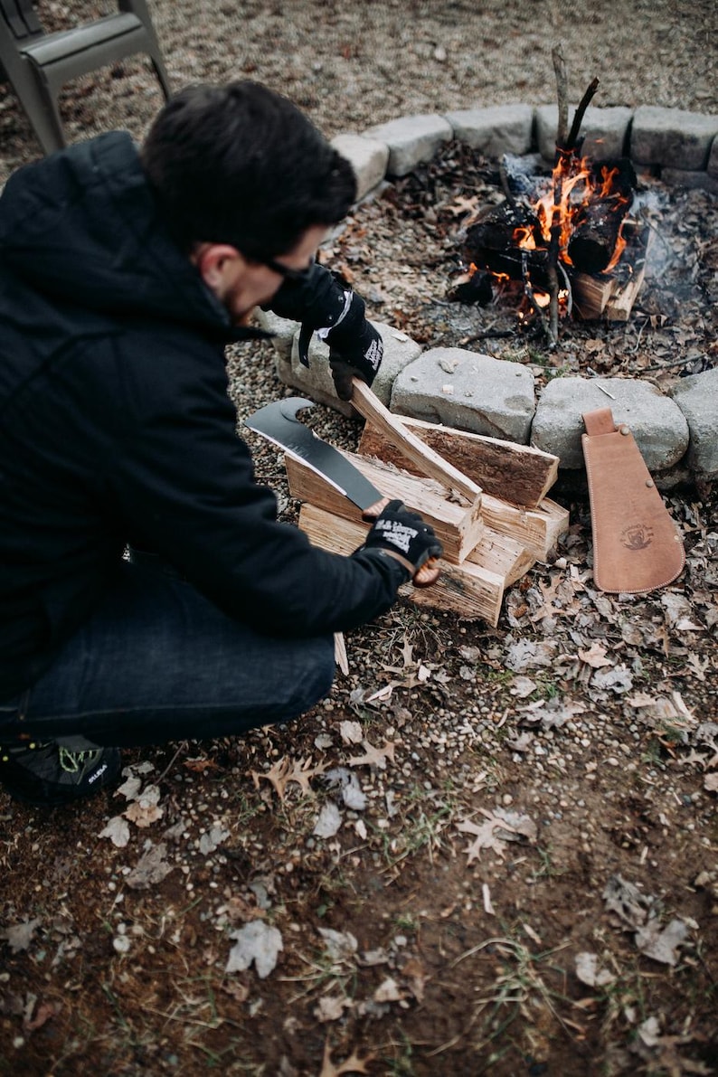 May include: A person wearing black gloves uses a large black knife to split a piece of firewood near a campfire. The knife has a curved blade and a leather sheath.