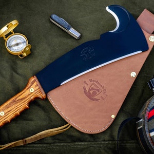 May include: A black and brown Woodman's Pal axe with a leather sheath and a wooden handle. The axe has a curved blade and a small hole in the top of the blade. The sheath has a brown leather strap and a brass buckle. The axe is on a green canvas background.