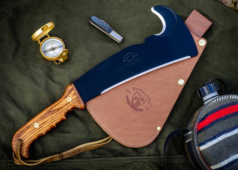 May include: A black and brown Woodman's Pal axe with a leather sheath and a wooden handle. The axe has a curved blade and a small hole in the top of the blade. The sheath has a brown leather strap and a brass buckle. The axe is on a green canvas background.