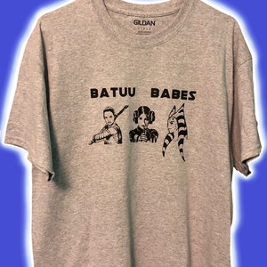 May include: A gray t-shirt with the text "BATUU BABES" printed on the front. The shirt features three black and white illustrations of female characters from the Star Wars franchise.