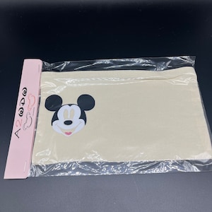 May include: A cream-colored canvas pouch featuring a black and white Mickey Mouse graphic. The pouch is sealed in clear plastic with a black zipper closure. A pink tag with text is attached to the side.