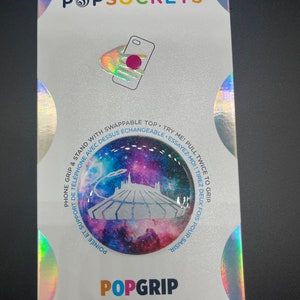 May include: A white PopSockets PopGrip phone grip with a galaxy-themed design. The design features a white silhouette of a castle against a purple, pink, and blue galaxy background. The text "TRY ME! PULL TWICE TO GRIP" is printed in blue on the white background. The text "POINÉE ET SUPPORT DE TÉLÉPHONE AVEC DESSUS ÉCHANGEABLE - ESSAYEZ MOI! TIREZ DEUX FOIS POUR SAISIR" is printed in blue on the white background.