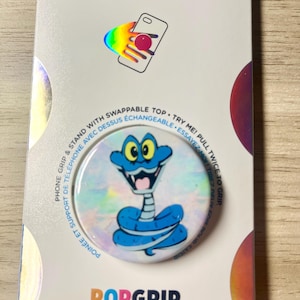 May include: A PopSockets PopGrip featuring a blue cartoon snake design. The PopGrip is on a white background with the words "POPSOCKETS" and "POPGRIP" printed on the packaging. The packaging also includes the text "PHONE GRIP & STAND WITH SWAPPABLE TOP. TRY ME! PULL TWICE TO GRIP."