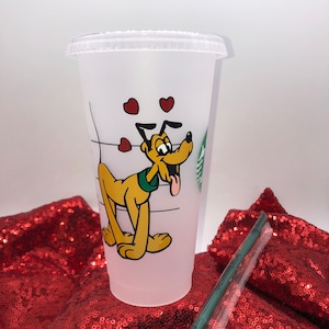 May include: A white reusable cup with a cartoon image of Pluto the dog with red hearts. The cup is on a red sparkly surface.