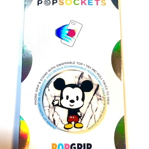 May include: A PopSockets PopGrip featuring Mickey Mouse. The PopGrip has a white marble-like background with a cartoon image of Mickey Mouse. The packaging includes the PopSockets logo and text that reads "Phone Grip & Stand with Swappable Top."