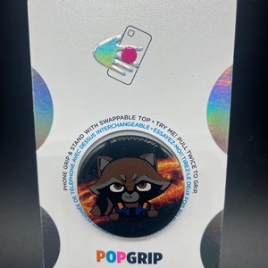 May include: A black and white PopGrip phone stand with a cartoon image of Rocket Raccoon from Guardians of the Galaxy. The background is a galaxy with a swirling nebula. The text "PopGrip" is printed in white on the bottom of the stand.