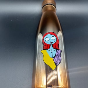 Op de afbeelding: Een roestvrijstalen waterfles met een verloop van zwart naar goud. De fles heeft een bedrukt beeld van Sally uit The Nightmare Before Christmas.