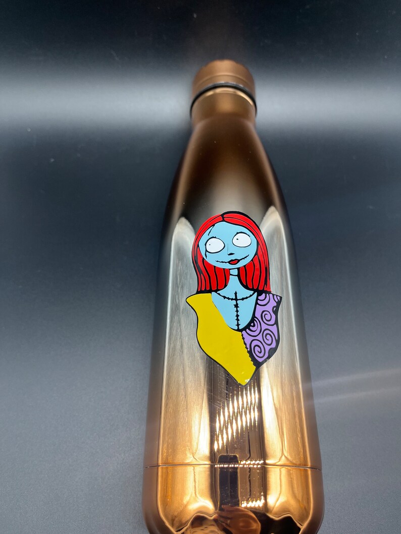 Op de afbeelding: Een roestvrijstalen waterfles met een verloop van zwart naar goud. De fles heeft een bedrukt beeld van Sally uit The Nightmare Before Christmas.