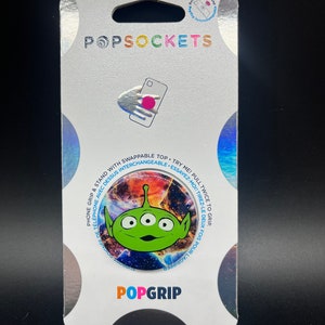 May include: A green alien character with three eyes from the movie Toy Story is featured on a phone grip. The phone grip is a round, expandable stand with a colorful galaxy background. The packaging is white with the words "PopSockets" and "PopGrip" printed on it.