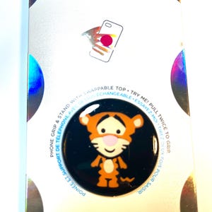 May include: A PopSockets PopGrip featuring Tigger from Winnie the Pooh. The black PopGrip has a glossy finish with a cartoon image of Tigger in orange and yellow. The packaging reads "POPSOCKETS" and "POPGRIP".